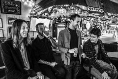 Four band members laughing, drinking, and sitting at a bar.